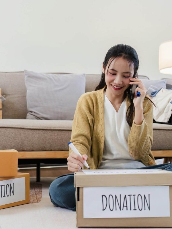 Young woman Asian of charity putting clothes in boxes with donations lettering and using smartphone